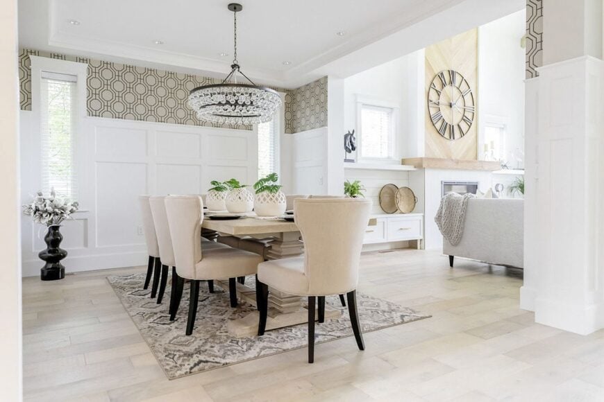 The dining room offers a patterned rug, beige wingback chairs, and a light wood dining table illuminated by a round chandelier.