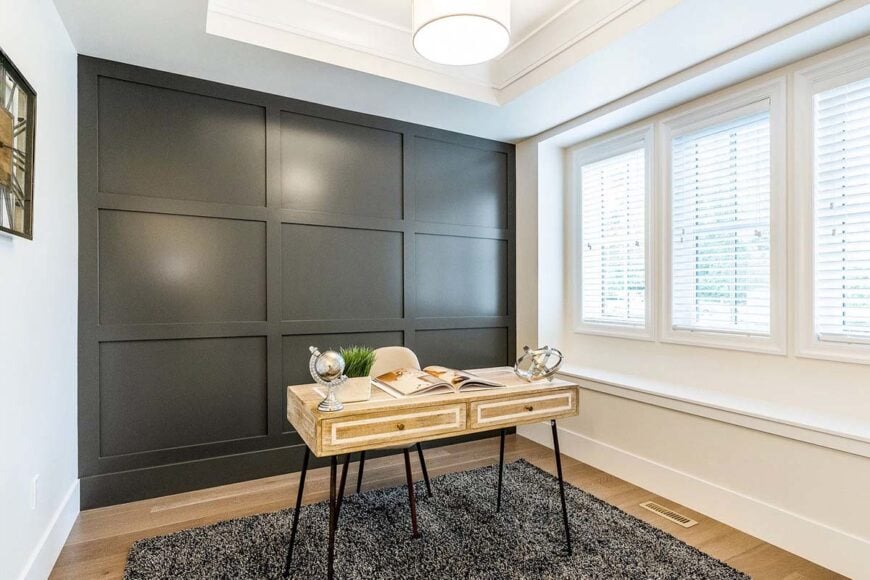 Study with a tray ceiling, paneled accent wall, louvered windows, and a wooden desk that sits on a gray shaggy rug.