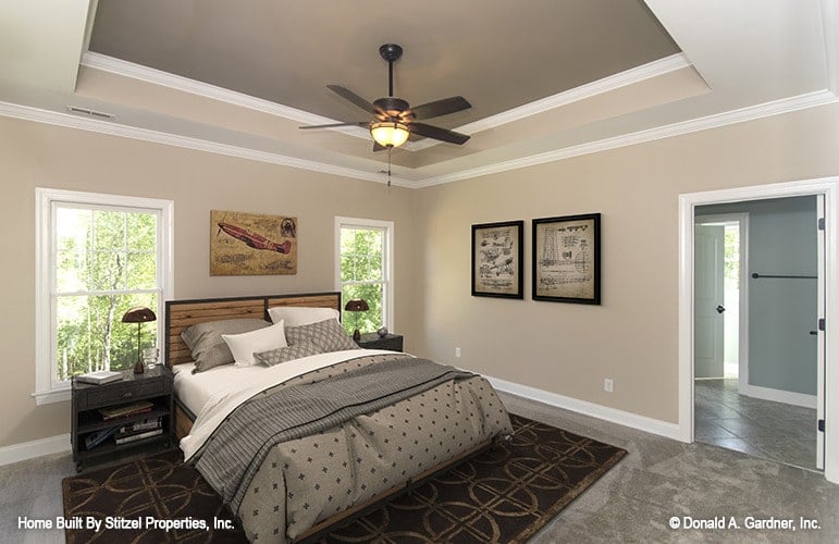 Primary bedroom with a tray ceiling, cozy wooden bed, and beige walls adorned with various artworks and white moldings.