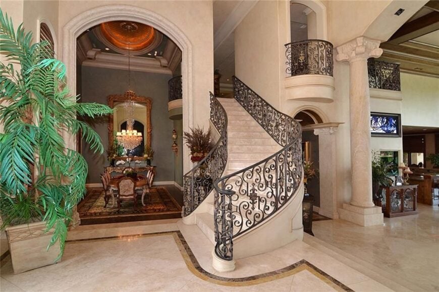 Foyer with a winding staircase framed with ornate wrought iron railings. An archway beside it opens to the formal dining room.