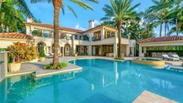 This is a look at the back of the mansion with a large swimming pool adorned by tall palm trees that bring color to the beige exteriors of the house with large windows. Image courtesy of Toptenrealestatedeals.com.