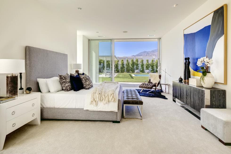 The gray frame of bed in this bedroom pairs well with the bright beige walls and ceiling. This si then complemented by bright bedside drawers and the glass wall at the far end. Image courtesy of Toptenrealestatedeals.com.