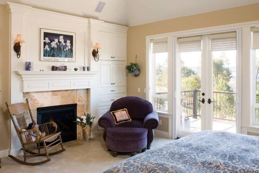 The primary bedroom has a cozy bed, a purple lounge chair, a wooden rocking chair, a fireplace, and a private deck.