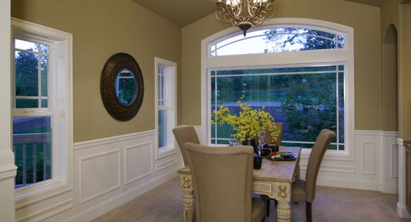 The dining room has a rustic table, high back upholstered chairs, and beige walls adorned with a round mirror and white wainscoting.