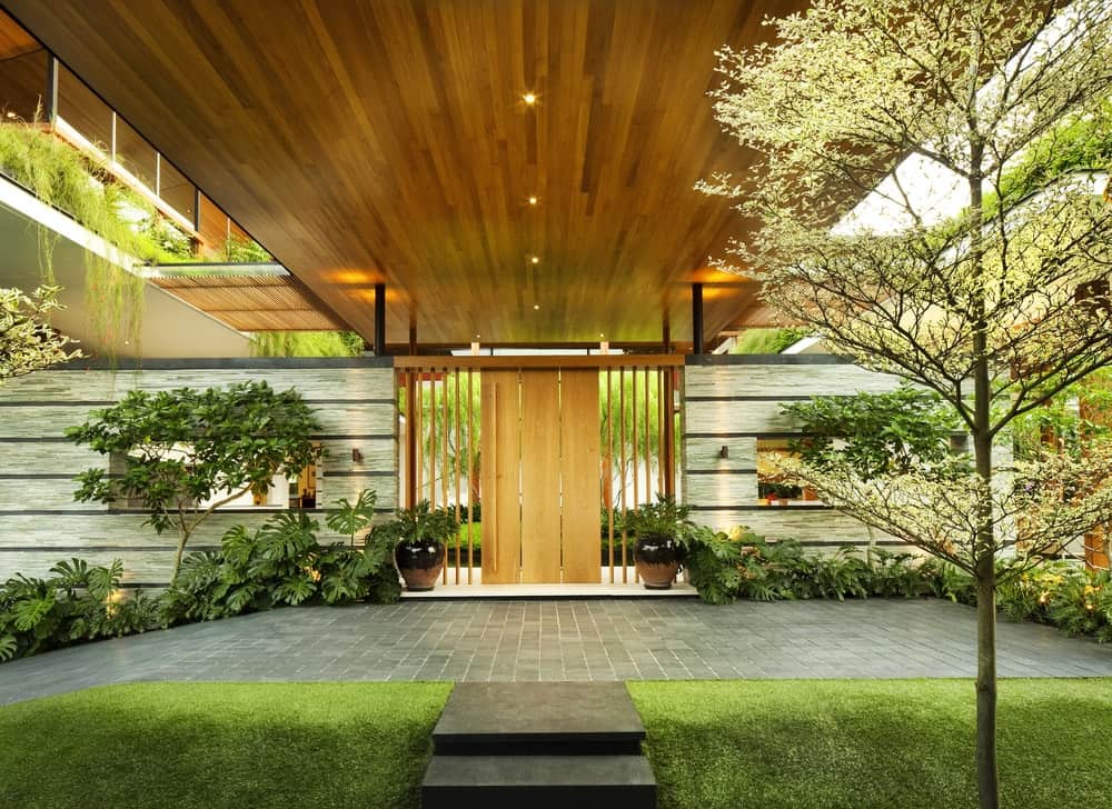 This is the view of the outer entrance to the courtyard. the wooden door matches well with the ceiling above with recessed lights. These are then complemented by lush landscaping of shrubs.