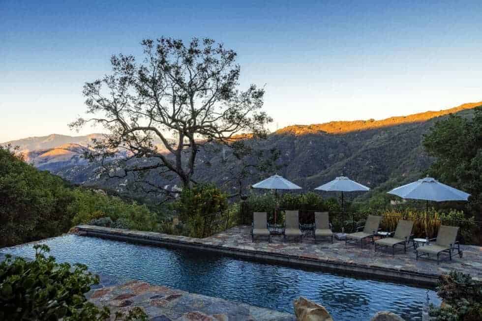This is a view of the pool that is built on a cliff with a sweeping view of the valley below. The pool is also bordered with stone walkways and shrubs. Image courtesy of Toptenrealestatedeals.com.