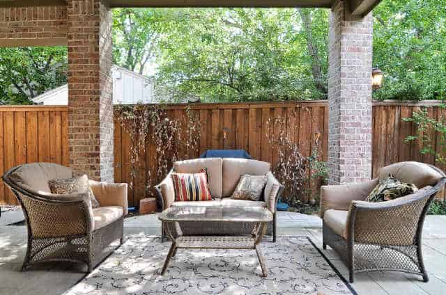 Covered patio with wicker cushioned chairs and a glass top coffee table that sits on a patterned rug.