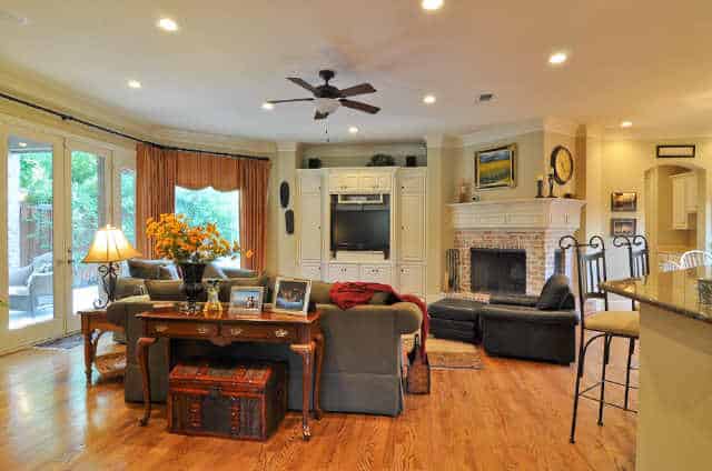 The family room has a corner fireplace, a TV, gray seats, wooden tables, and a french door that leads out to the veranda.
