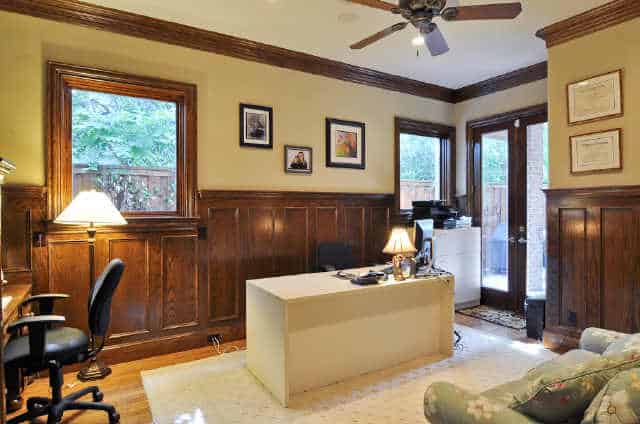 The study has built-in and white center desks, swivel chairs, and yellow walls adorned with framed artworks and wooden wainscoting.
