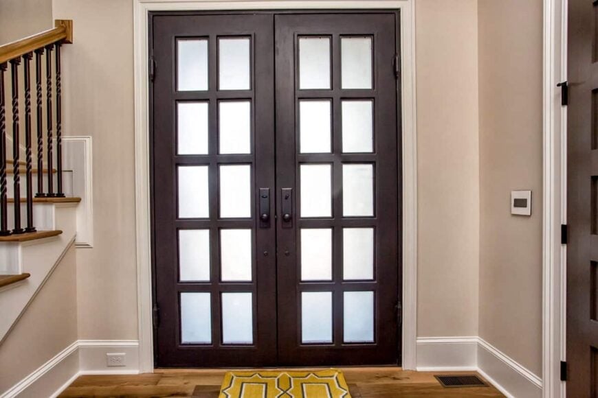 The french front door opens to the foyer with beige walls and hardwood flooring topped by a yellow patterned rug.