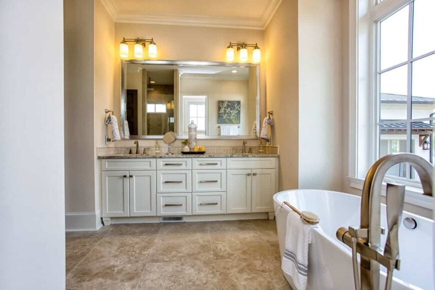Primary bathroom with dual sink vanity and a freestanding tub placed under the white framed window.