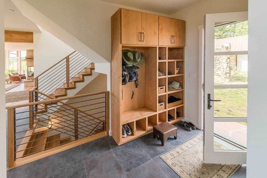 Mudroom with wooden cabinet and shelving, a brown stool, and a bordered rug that lays on the concrete tiled flooring.
