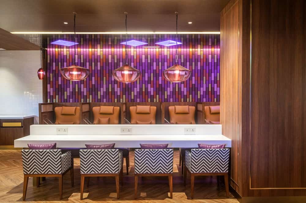 The long table with electrical sockets is topped with a row of pendant lights hanging from an earthy brown ceiling that pairs well with the wooden tones of the walls and the flooring.