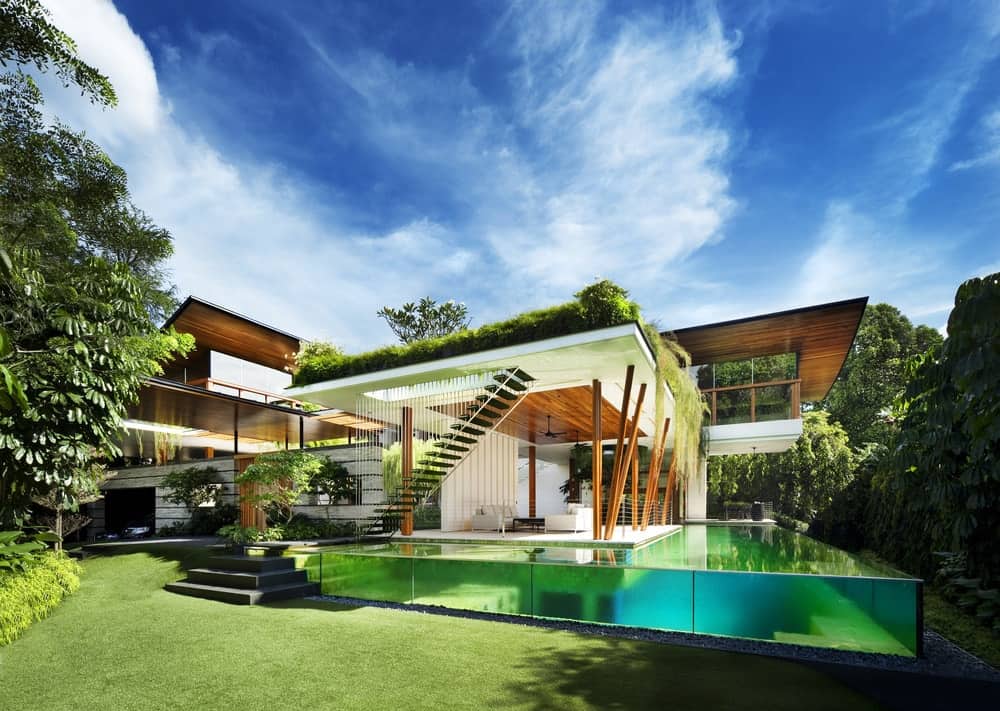 This is an exterior view of the side of the house showcasing the glass walls of the infinity-edge pool attached to the house that has a lot of outdoor areas with open walls.