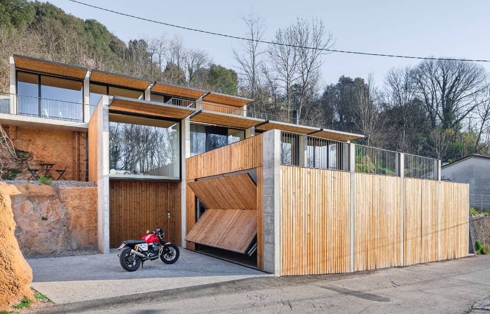 This is a view of a modern house with a fancy design. The walls are decorated with wooden planks and glass walls. There is also a garage with a foldable door.
