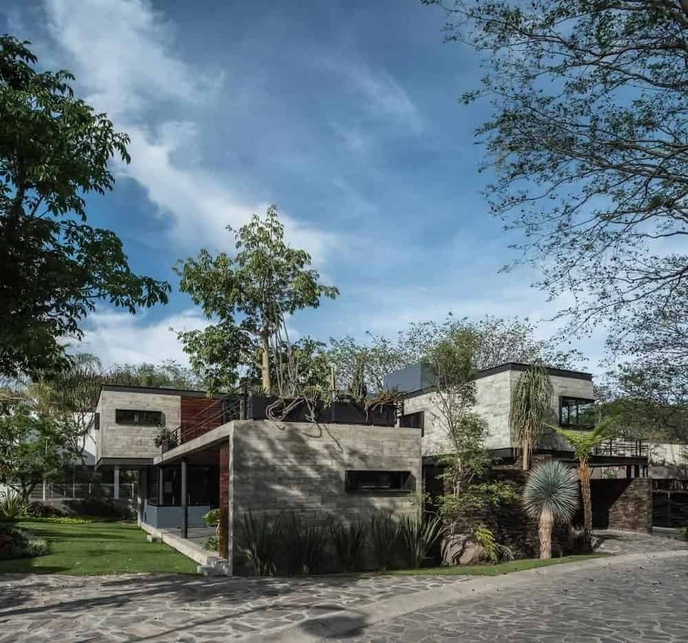 This is a look at the side of the house with mosaic stone walkways and driveways to match with the concrete bricks of the exterior walls of the house adorned with windows, glass walls and lush landscaping.