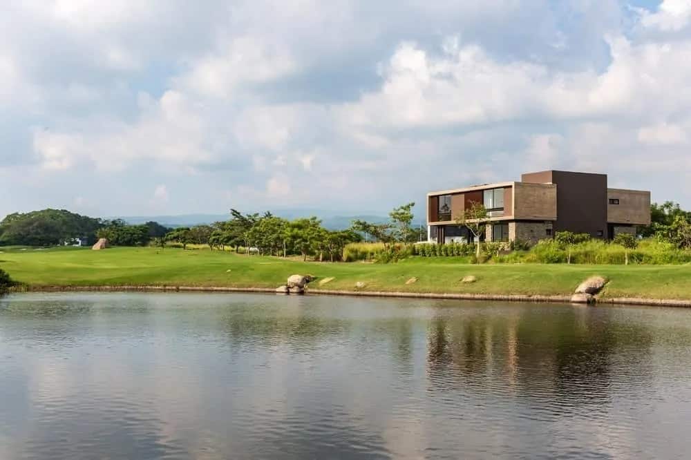 This is a look at the back of the house with earthy exterior wall tones that make it stand out against the surrounding green landscape with a large pond.