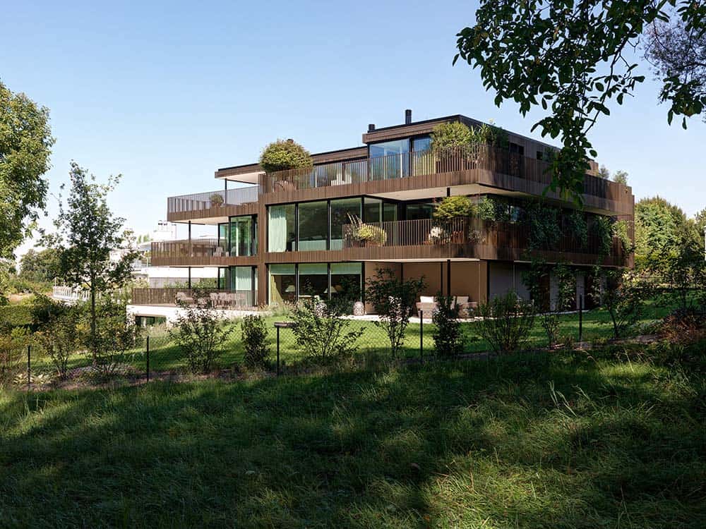 This is a view of the house from the back showcasing its dark wooden exterior with an abundance of glass walls and balconies complemented by various potted plants and the landscaping.