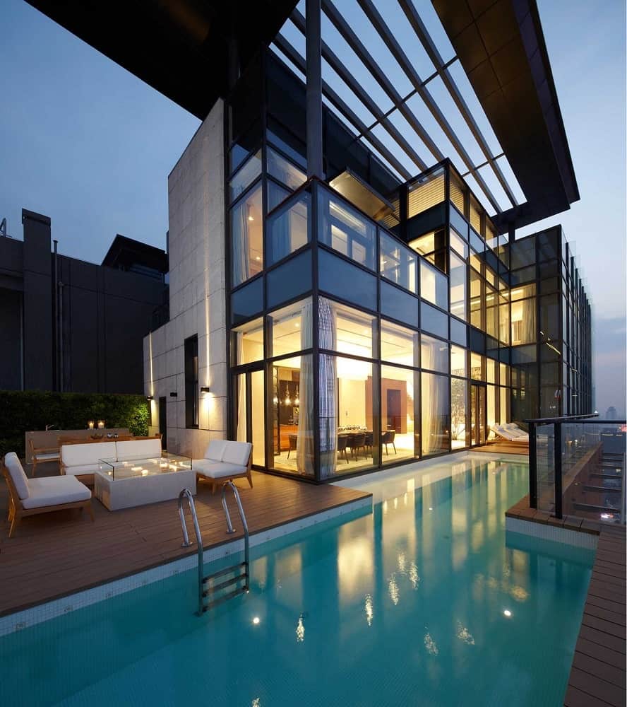 This is a view of the house from the vantage of the poolside area. You can see here that the house is mostly made of glass walls that glow warmly from the interior lights that escape to the wooden deck walkways outside.