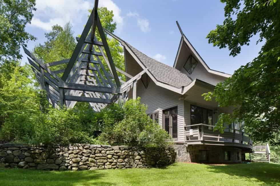 This is a view of the back of the house that has a unique tall A-frame roof design paired with shiplap exterior walls and lush landscaping of grass, trees and shrubs.