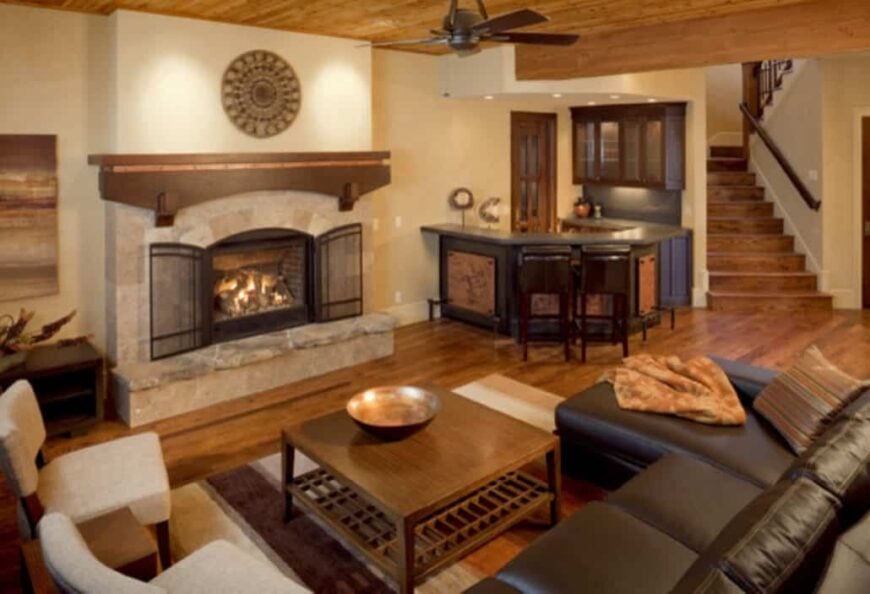 Recreation room with L-shaped sectional, beige chairs, wooden coffee table, a fireplace, and a wet bar.