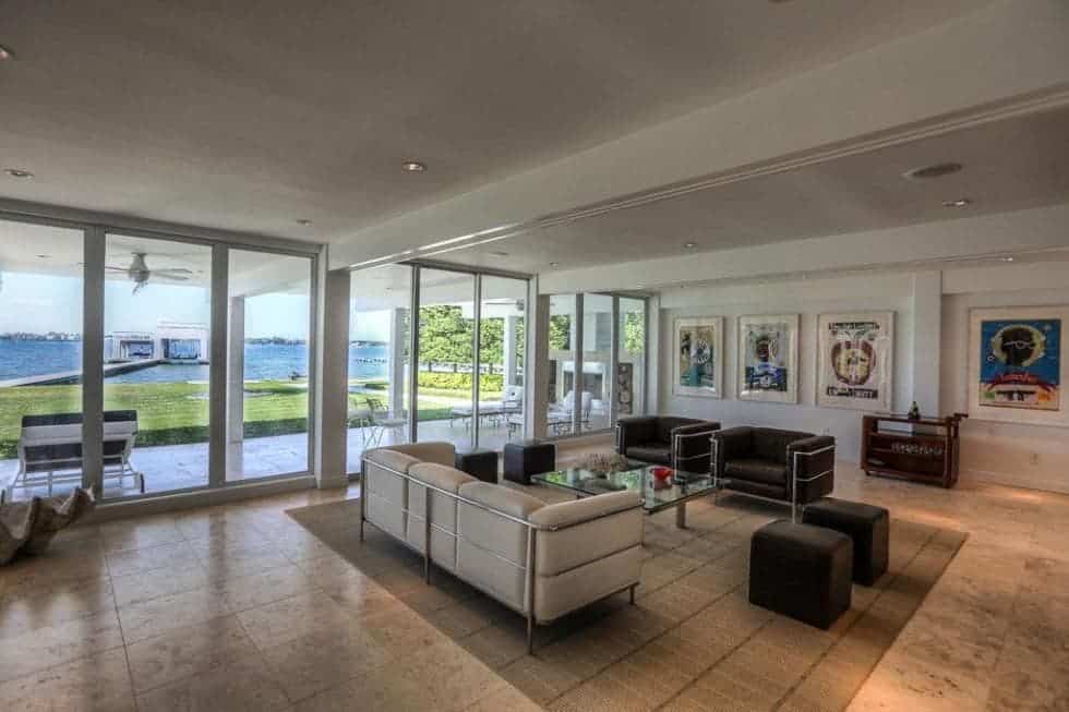 This is the living room that has a beige sofa paired with dark brown armchairs and ottomans surrounding a glass-top coffee table. These are brightened by the bright ceiling and glass wall. Image courtesy of Toptenrealestatedeals.com.