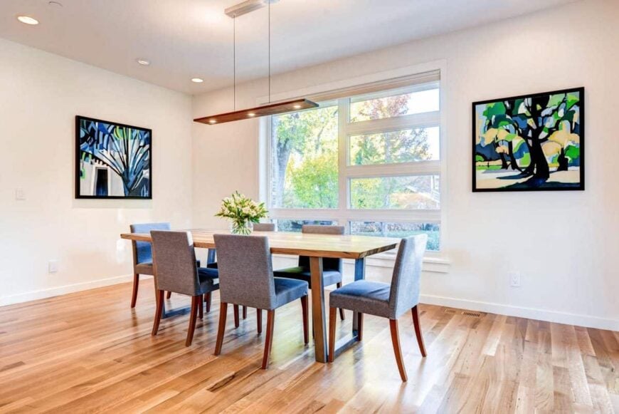 The dining area offers a linear pendant, rectangular dining table, gray upholstered chairs, and bold paintings adorning the white walls.