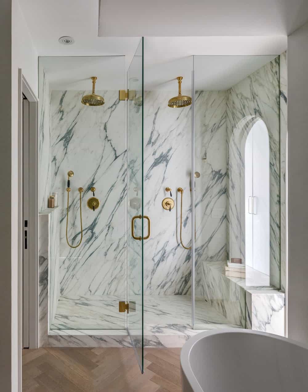 This is a look at the glass-enclosed shower area of the bathroom with white marble walls and floor that makes the bronze fixtures stand out.