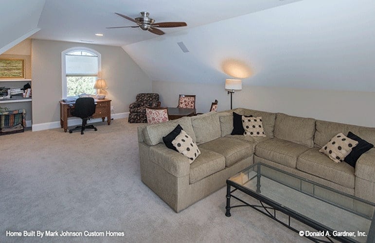 The bonus room is filled with a glass top table, beige sectional, small dining set, and a desk placed under the arched window.