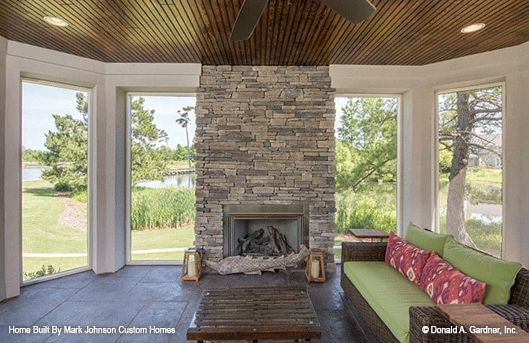 The sunroom has a large stone fireplace, wooden coffee table, and wicker seats topped with green cushions and patterned pillows.