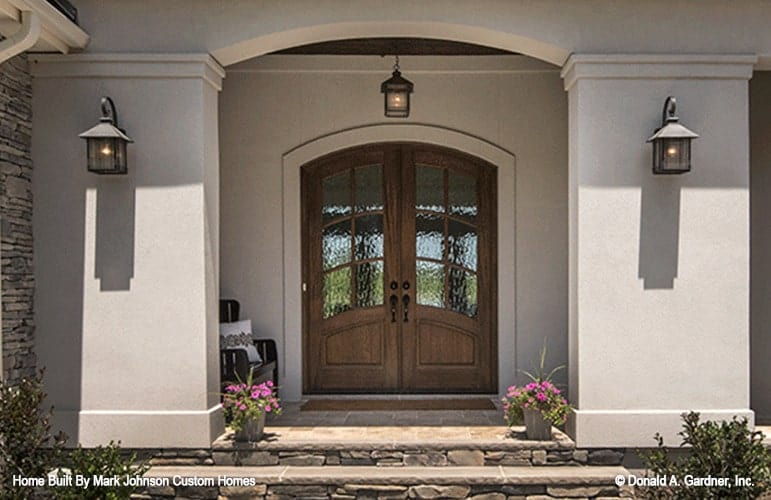 A closeup view of the home entry with an arched french front door well-lit by a glass pendant and matching sconces.