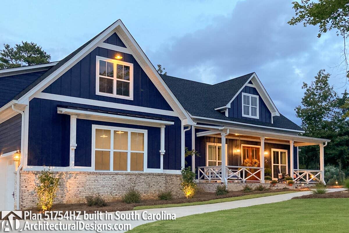 Alternative front exterior with dark blue vertical siding, white-framed windows, stone bases, and a covered porch enclosed in white railings that are arranged in a crisscross pattern.