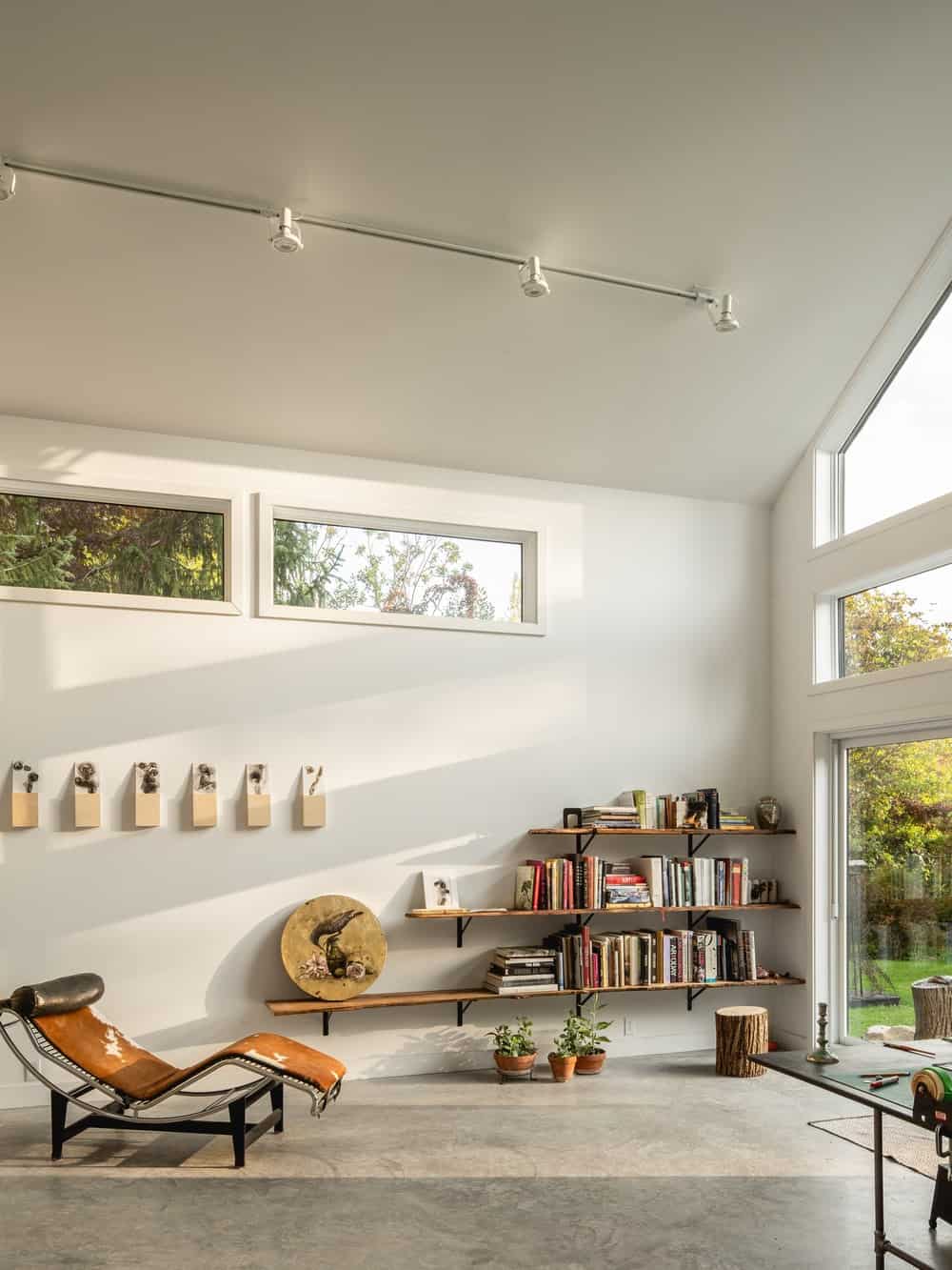 At the back of the extension is a large door that leads to the backyard by the large glass wall that illuminates the wall of shelves on the side with a lounge chair.