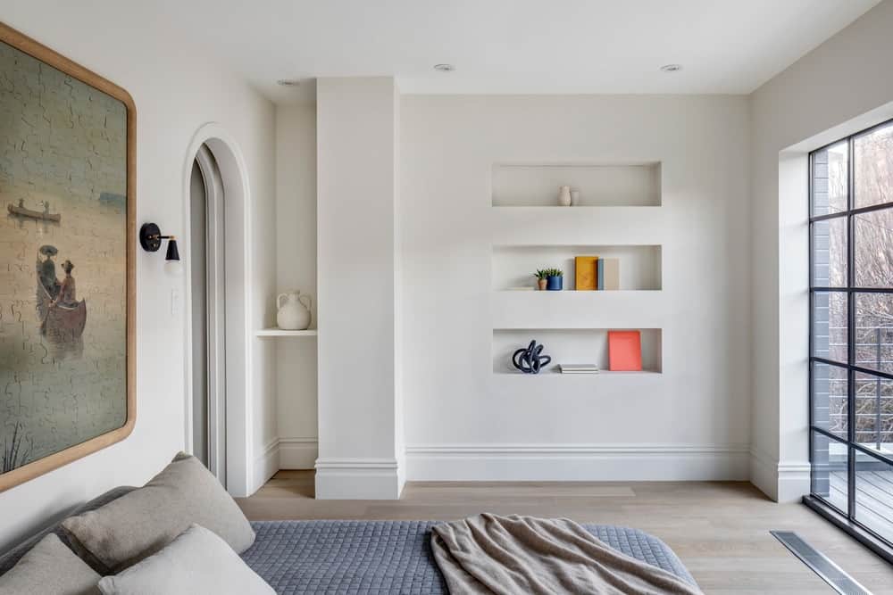 This is the simple bedroom with a cottage bed topped with a large painting and an opposite bright wall with built-in shelves brightened by the French-style glass door.