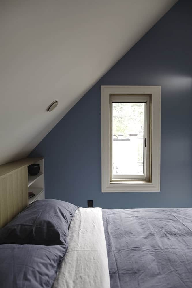 The bedroom has a shed ceilingand gray wall with a window that brings in natural lighting for the gray bed.