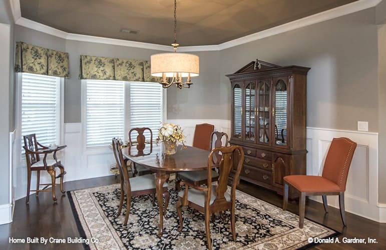 Formal dining room with a wooden display cabinet, an oval dining set, floral area rug, and a bay window dressed in patterned valances.