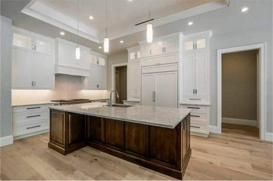 The kitchen has a tray ceiling, white cabinetry, and an immense prep island well-lit by cylindrical pendants.