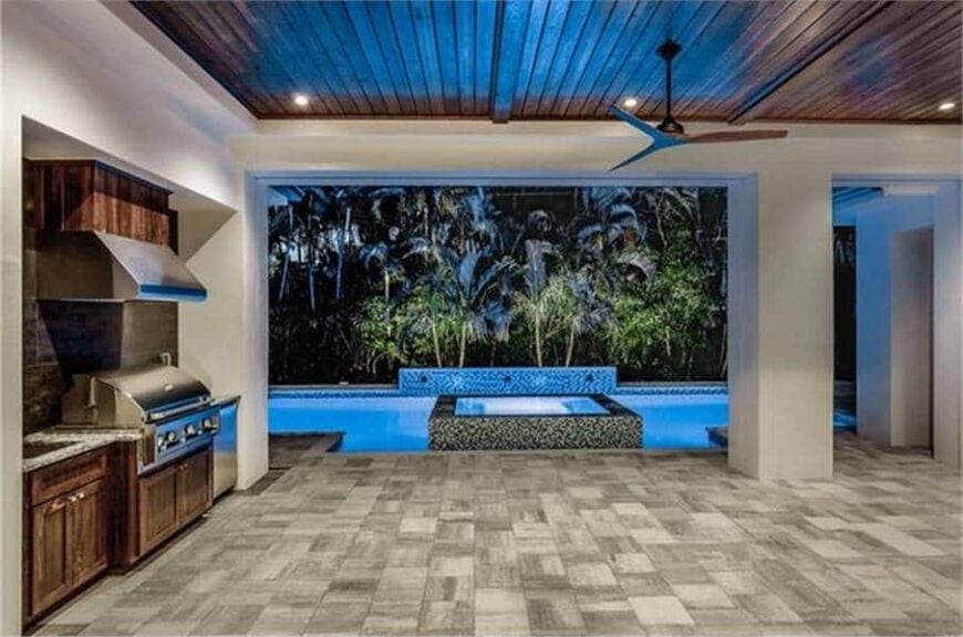 The covered patio has a summer kitchen and a fan hanging from the tray ceiling clad in wood planks.