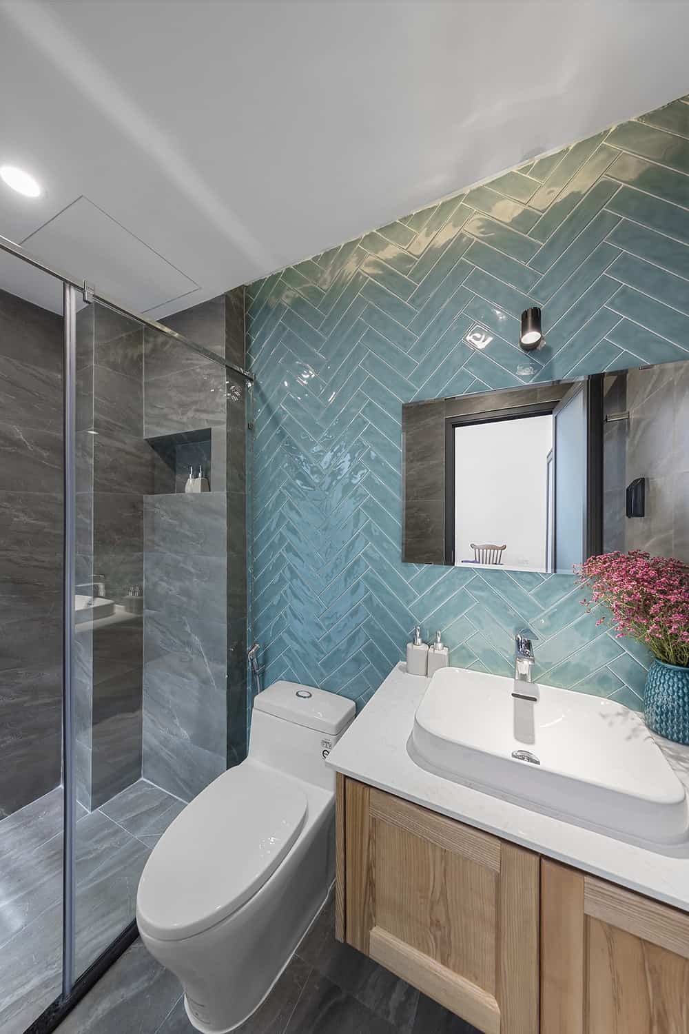 The bathroom has a light blue herringbone wall that contrasts the white porcelain toilet and sink. On the far side is the glass-enclosed shower area with dark gray tones,