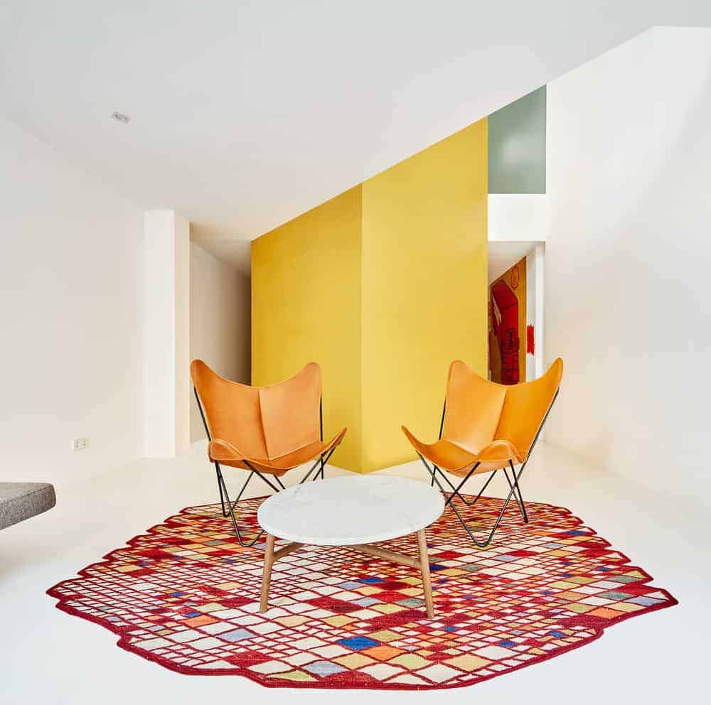 This is a close look at the living room with a couple of orange chairs on a red patterned area rug that makes it stand out against the white floor.