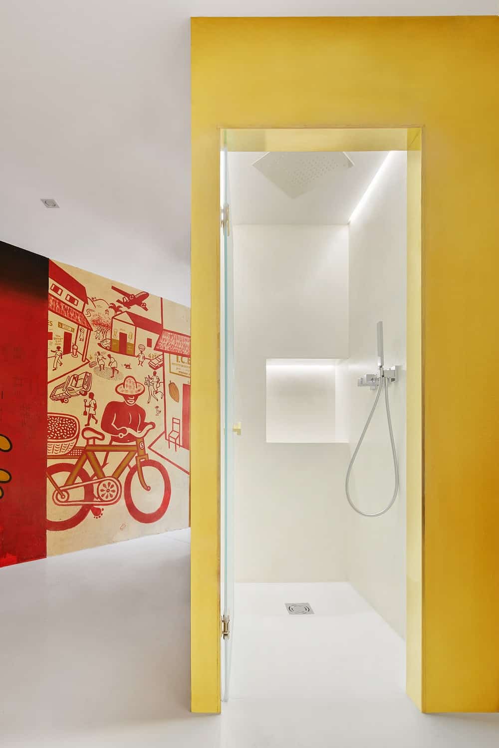 A few steps from the detailed mural wall is the shower area that has bright walls and flooring behind a glass door.