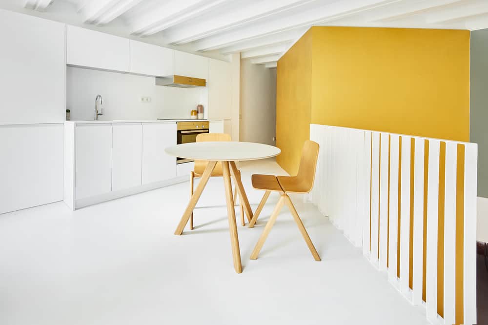 A few steps from the cooking area of the simple kitchen is a small round table for a breakfast nook by the indoor balcony with white railings.