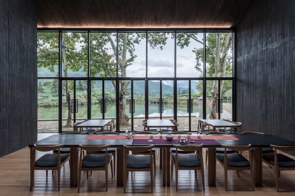 This large room has multiple dining tables with a dark tone that matches the walls and balanced by the natural lighting that goes in from the glass walls and door on the far side.