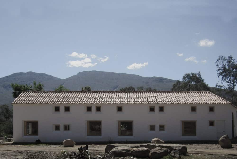 This is a full view of the house with adobe walls, red clay tiles on the roof and multiple small and large windows that stand out against the bright tone of the exterior walls.