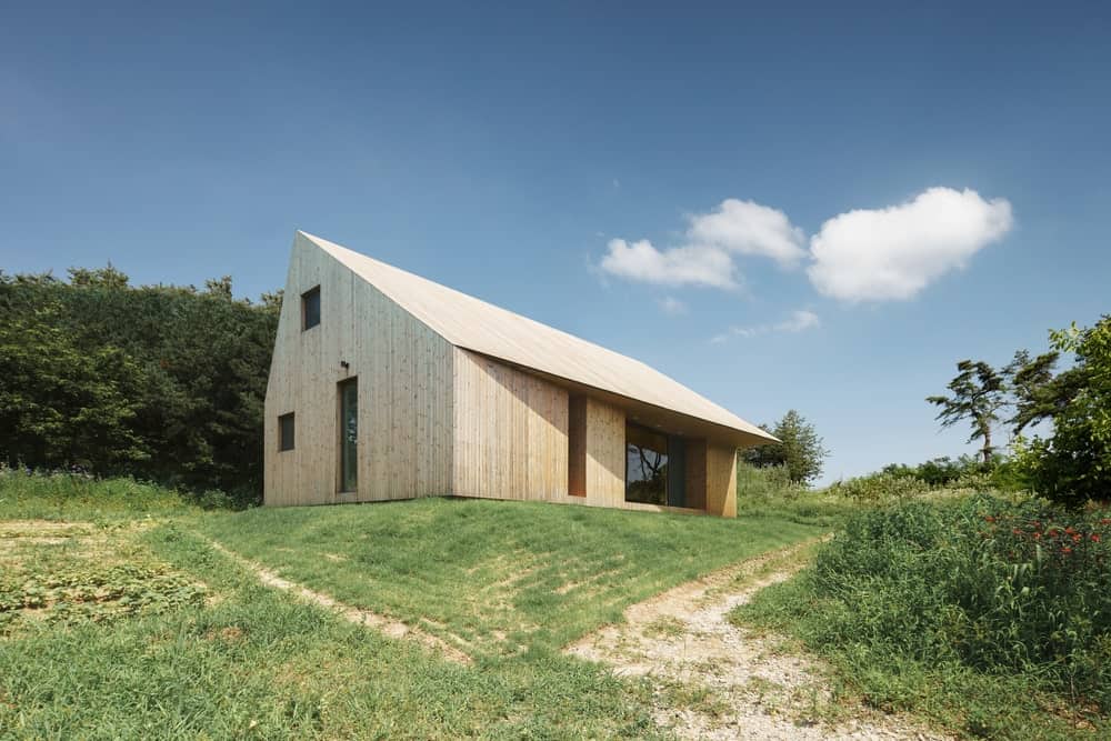 This is an exterior look at the house with a simple earthy wooden tone that pairs well with the surrounding grass lawn and tall trees.
