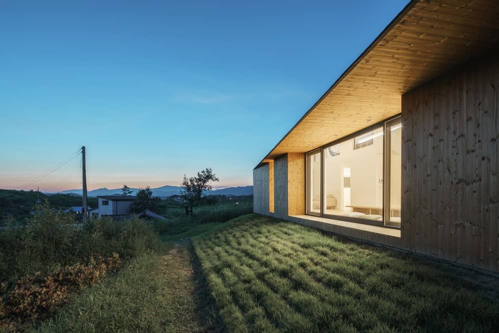 This is a close look at the exterior of the house with a large glass wall that glows warmly and shows the glimpse of the interior.