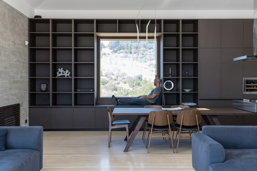 By the long rectangular wooden table of the dining area is a reading nook by the window surrounded by bookshelves.