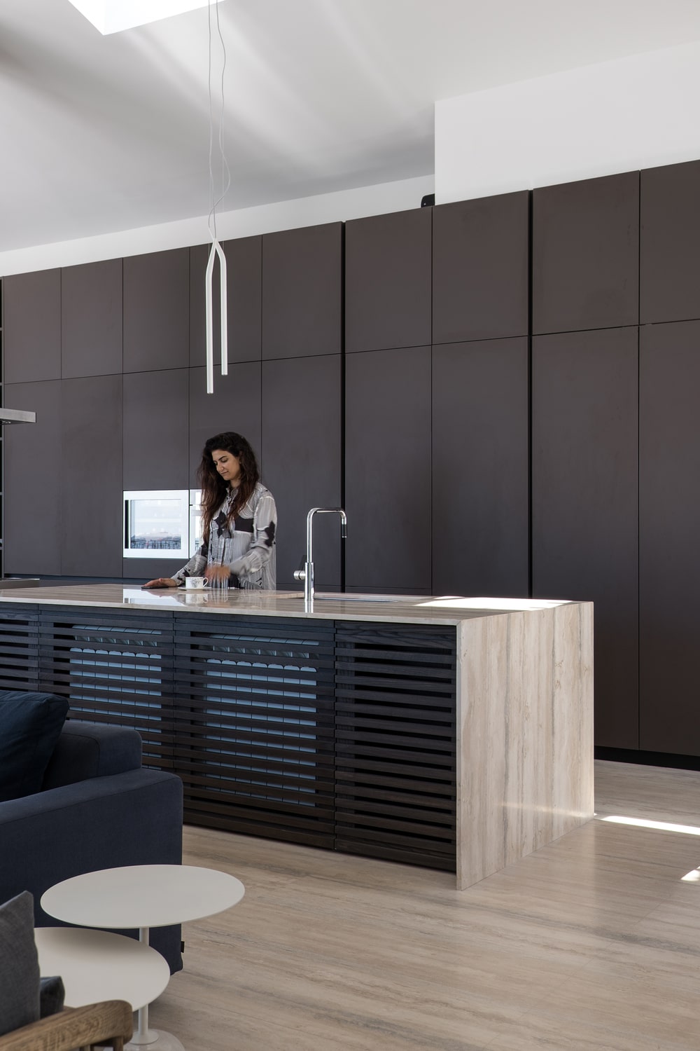 The kitchen has a large waterfall island with a tone to match the floor and contrast the dark modern cabinetry on the wall.