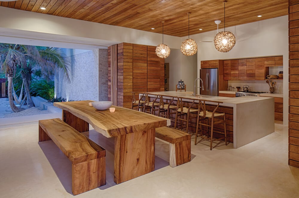 A few steps from the living room sofa is the wooden dining table and the large kitchen island topped with spherical pendant lights.