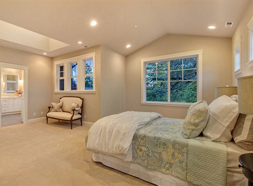 Primary bedroom with carpet flooring, vaulted ceiling, white-framed windows, a sitting area, and a comfy skirted bed.